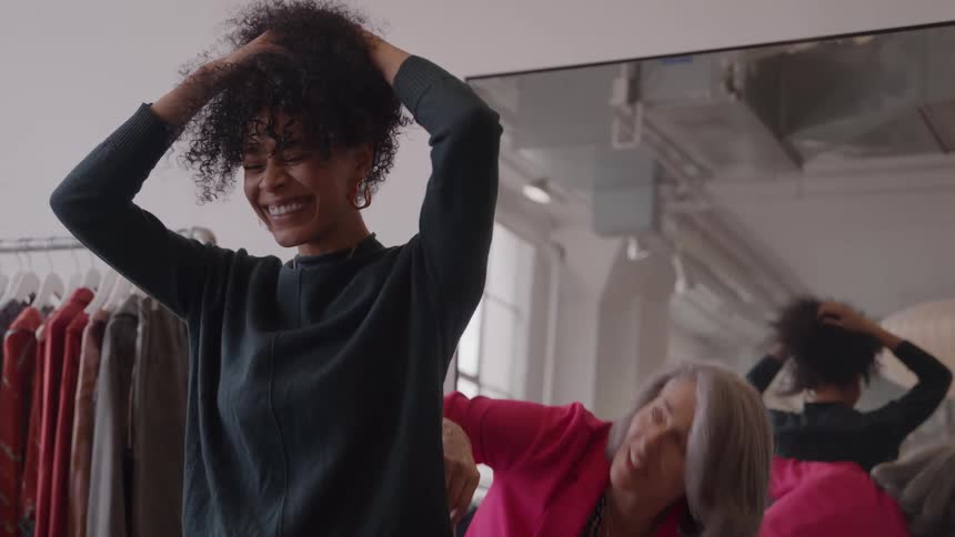 A curlyhaired woman with a large smile stands with hands in her hair while a second woman in a pink blazer stands slightly behind her measuring her clothing with a large mirror reflecting the scene on the right