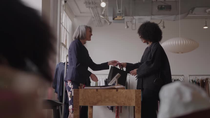 The video clip is set inside a brightly lit boutique featuring two women at a checkout counterthe older woman possibly an employee is giving a shopping bag to the younger woman with curly hair who smiles happily as she completes her purchase