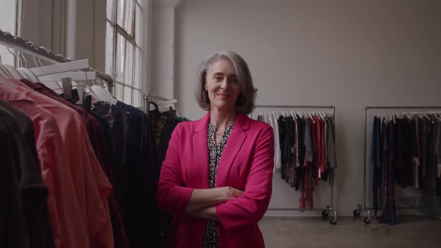 A woman with gray hair and a pink blazer stands in the center of a retail store with clothing racks on both sides and a large window on the left facing the camera with arms folded and a slight smile