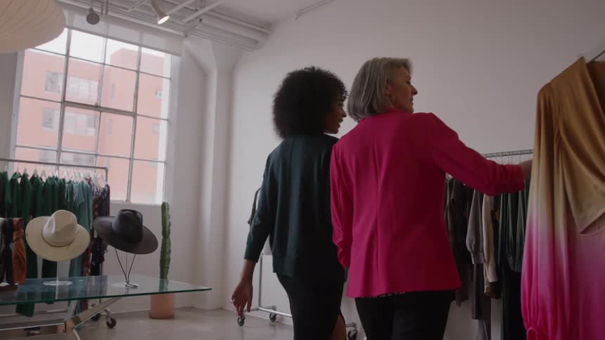 A grayhaired woman in a bright pink blazer and an afrohaired woman in a dark shirt look through a rack of clothes in a bright stylish clothing boutique with large industrial windows