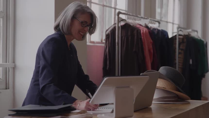 A happy grayhaired woman in a dark navy blazer and glasses smiles while working on a tablet pointofsale system behind a counter in a small clothing boutique with a rack of garments in the background
