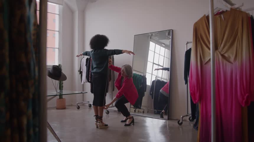 A woman in a red jacket and dark skirt kneels down next to a second woman with curly hair who stands with arms outstretched in front of a large mirror as the woman in the jacket appears to be adjusting the fit of the shirt on the standing woman