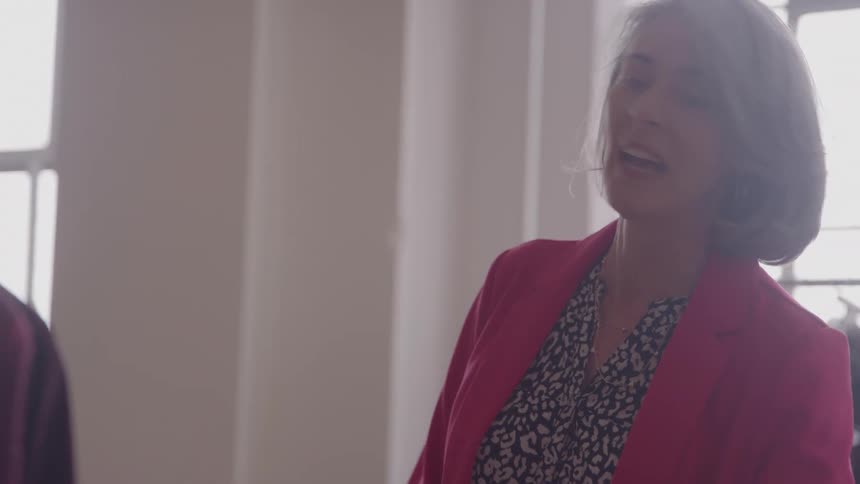 Grayhaired woman wearing a pink blazer and patterned blouse speaking in a bright room