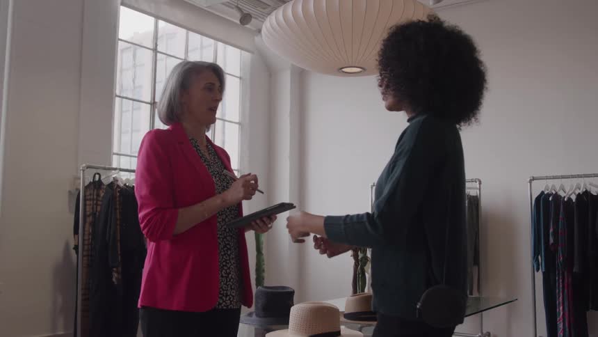 The video clip features two women in a brightly lit clothing store one older and one younger standing and chattingthe older woman wearing a pink blazer is holding a tablet or clipboard and appears to be discussing something businessrelated with th