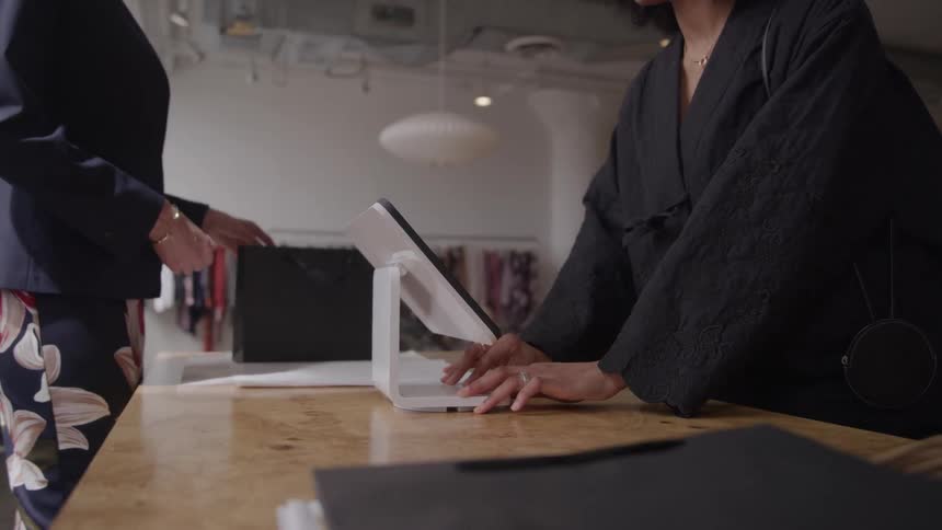 A woman stands inside a clothing shop at midday inserting her credit card into a payment processor she enters her details then turns the screen toward the cashier with a smile to complete her purchase