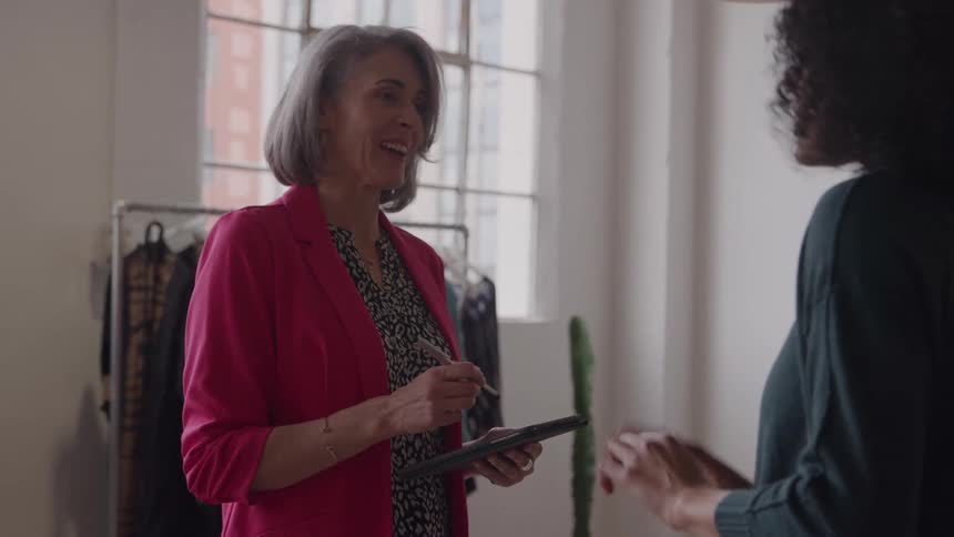 Grayhaired woman wearing a pink blazer and holding a tablet while talking to a client in a fashion studio