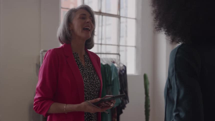 The video clip features an older woman with short gray hair and a bright pink blazer talking animatedly to a younger womanthey are standing in a boutique or clothing store and the older woman appears to be the sales associate or owner giving advi