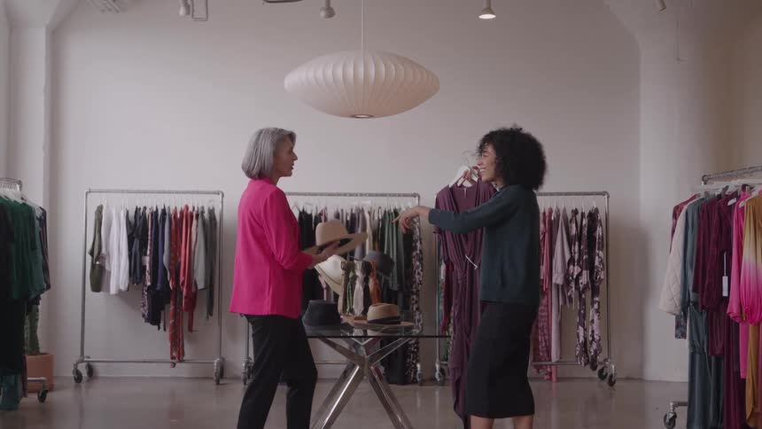 Two women in a boutique chat happily while comparing a hat and a dress amid racks of colorful clothing