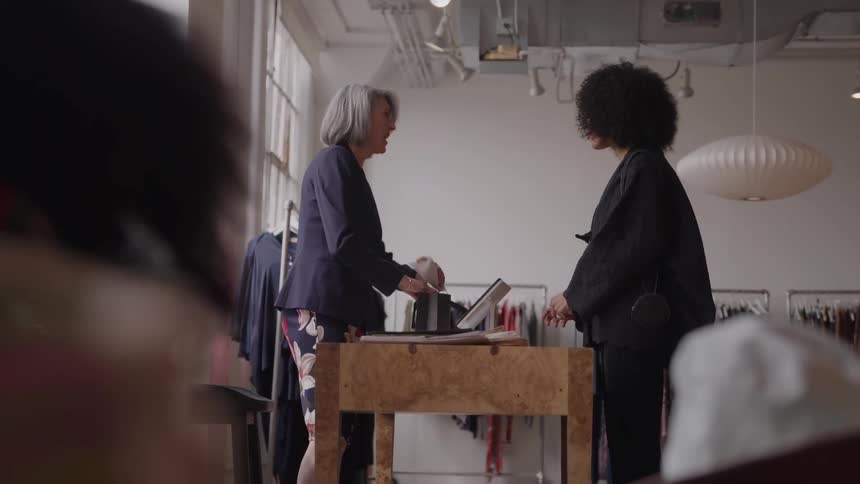 A female client stands in front of a female cashier inside a clothing store engaging in a brief discussion as the cashier bags the customers items with a friendly smile