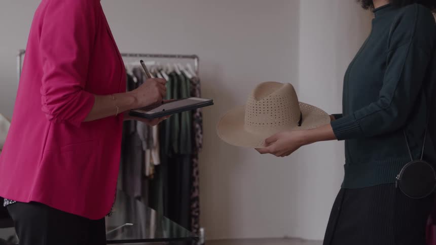 Woman in a pink blazer writing on a tablet while another holds a beige hat in a clothing store