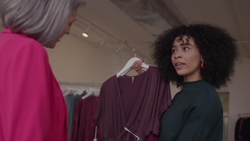 The video clip shows two women in a brightly lit clothing store having a conversationthe younger woman who has curly hair is holding up a dark longsleeved dress to the older woman who is wearing a pink blazer and they are both smiling
