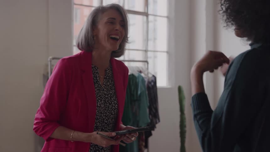 A woman in a pink blazer stands and laughs while holding a tablet facing and talking to a second woman who is seen from the back of the head and shoulder in a bright room with a large window and clothing racks