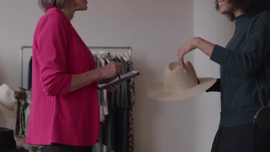 The video clip features two women in a brightly lit clothing store one older and one younger standing at a counterthe younger woman with curly hair is holding and examining a lightcolored straw hat