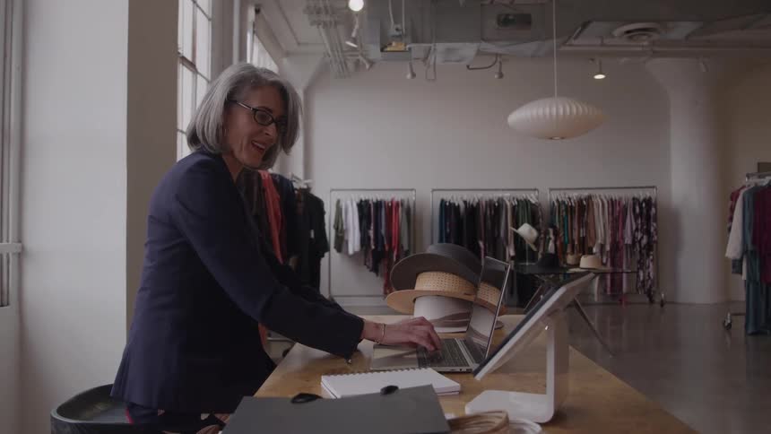 A stylish gray haired woman works on her laptop at the counter of a clothing boutique