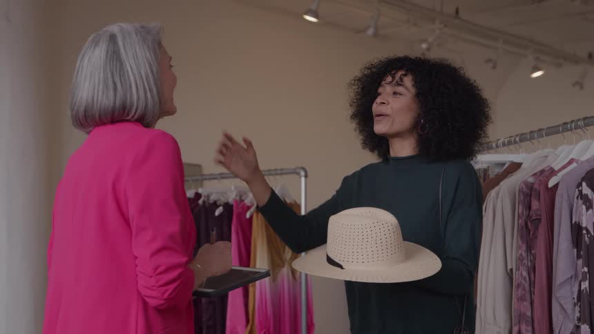 The video clip shows an older woman in a vibrant pink blazer and a younger woman with curly hair laughing and talking happily in a brightly lit clothing store the younger woman is holding a straw hat while engaging in the conversation with the ol