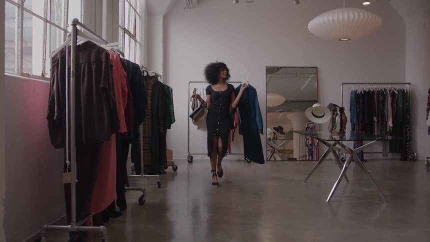 Woman in a black dress walks confidently through an airy boutique carrying shopping bags and a teal garment looking happy and selfassured