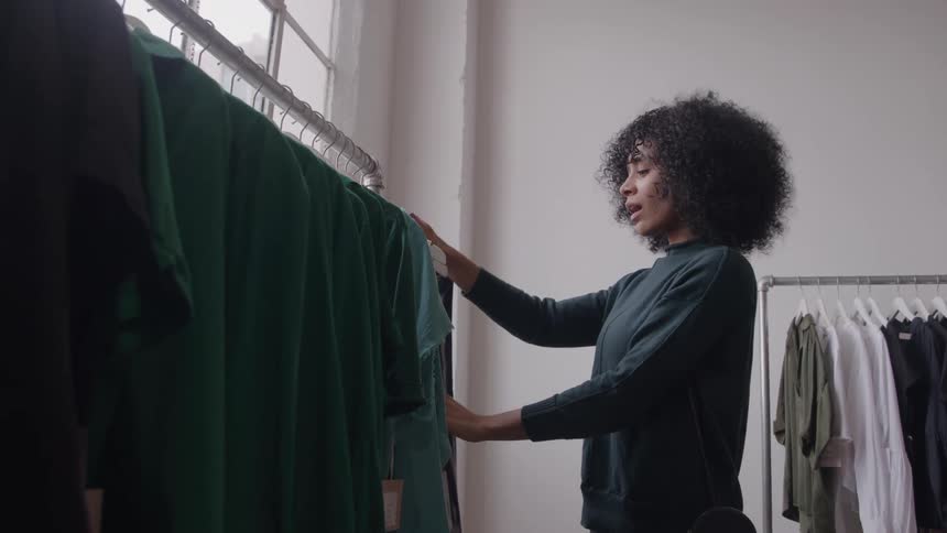 The video clip features a young woman with curly hair browsing a rack of dark green and black tops in a brightly lit clothing store she lightly touches the fabric of the garments as she examines the selection