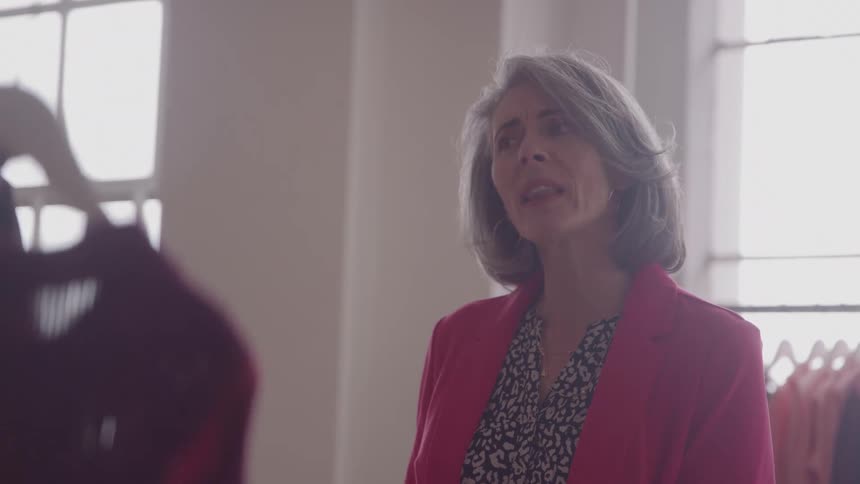 The video shows an older woman in a pink blazer looking up at a dark garment she is holding up to the light in a brightly lit clothing store she appears to be examining the quality or fit of the fabric