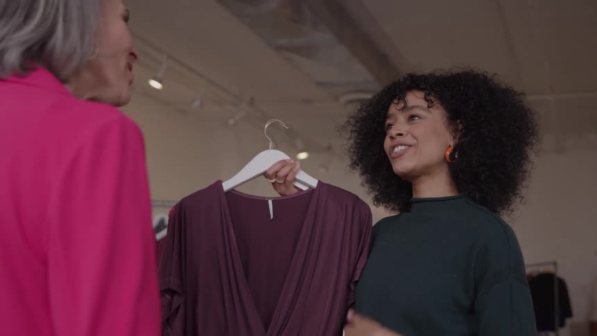 Curlyhaired woman excitedly discusses a mauve dress on a hanger with a stylist in a pink blazer