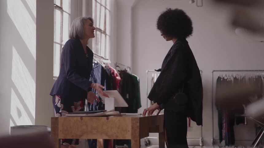 A grayhaired woman stands behind a wooden counter and hands a card reader to a second woman who is wearing dark clothing to make a payment in the middle of a bright retail space with large windows and clothing racks