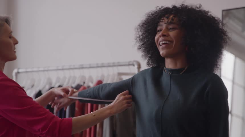 The video shows a saleswoman in a pink blazer helping a customer with curly hair inside a clothing store the saleswoman is measuring the customers arm with a tape measure while the customer smiles warmly indicating a positive shopping experienc