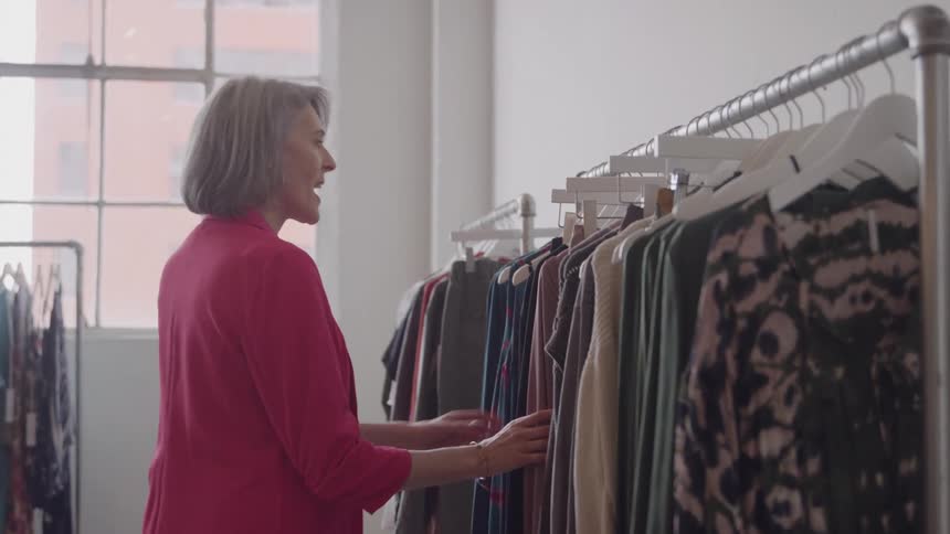A woman with gray hair and a red jacket stands next to a metal clothing rack and slides her hand along the hanging garments looking at the clothes in a bright room with a large industrial window