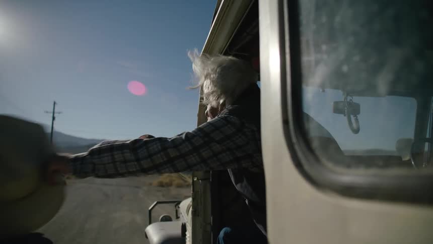 An elderly man tries to wave down a pickup truck from a moving vehicle