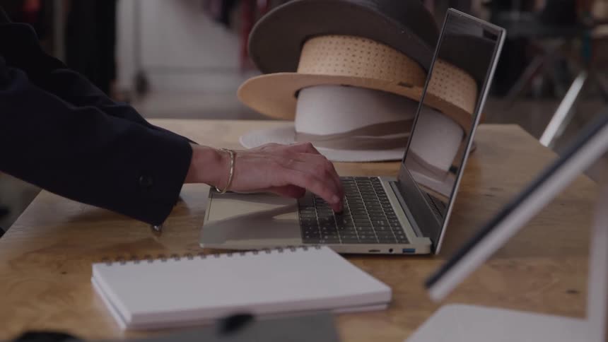Closeup of a persons hands typing on a laptop beside stacked hats and a small notebook on a wooden counter