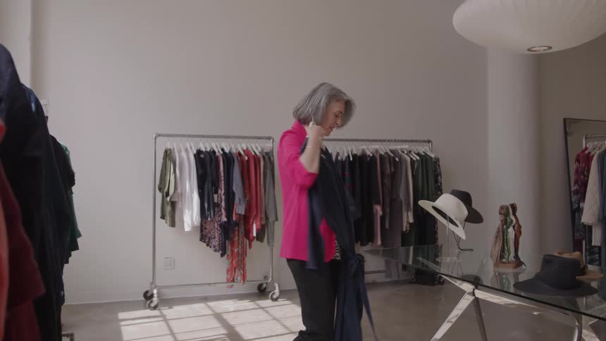 The video clip features an older woman in a bright pink blazer standing in a boutique she is holding up a dark possibly navy blue or black jumpsuit or dress for close examination