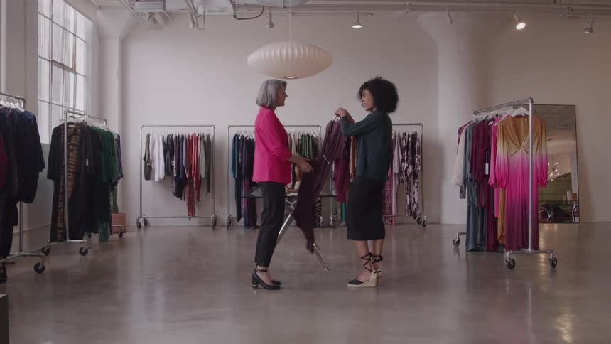 A grayhaired woman in a pink blazer stands facing a second woman in a dark skirt and top near a small table as the second woman holds up a dark piece of fabric and they converse in a bright open room lined with clothing racks