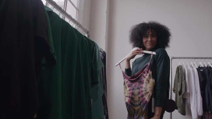 The video clip features a young woman with curly hair holding up a tiedye or patterned top to examine it in a clothing store she is also wearing a dark green sweater and is smiling slightly as she looks at the garment