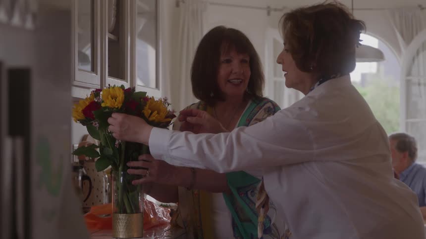 Two middleaged women one in a colorful printed top and the other in a white collared shirt happily arrange a large bouquet of red and yellow flowers in a vase in a brightly lit kitchen