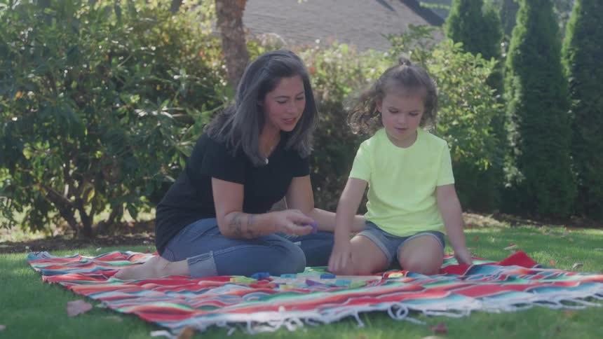 A woman and a young girl sit on a colorful blanket in the yard playing with pieces both focused and content