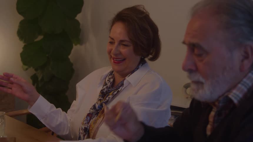The video features a warm closeup shot of an older woman and man sitting together at a table possibly diningthe woman is smiling broadly with her hands raised in a gesture of conversation while the man listens attentively next to her