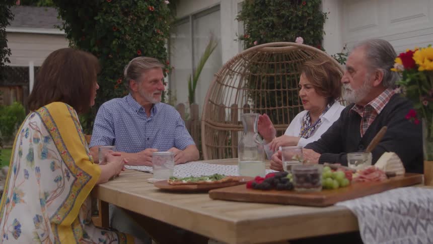 Two elderly men sit outdoors at midday with their wives beside them enjoying a relaxed lunch together a table in front of them holds fresh fruits and glasses of lemonade as they engage in cheerful conversation under the warm sun