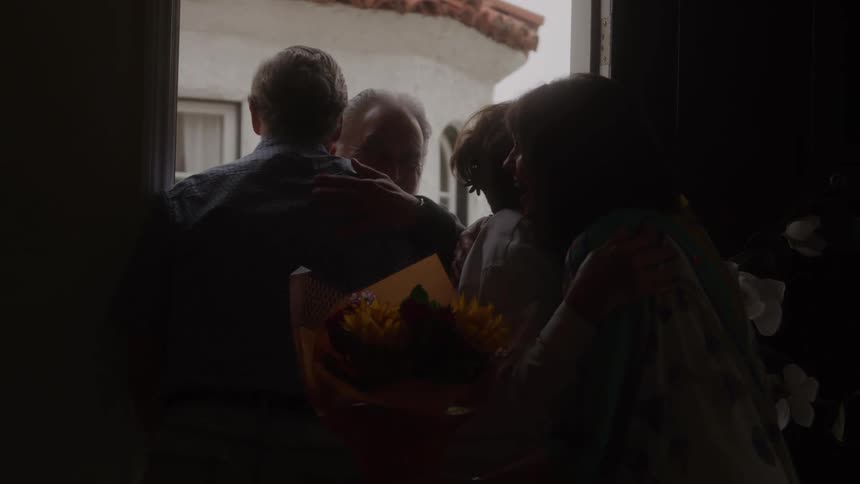 Two people welcoming an elderly couple in a doorway and one of the womans holding a bouquet and both couples embracing in a scene framed by the bright exterior