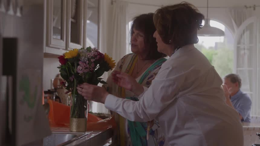 A medium view of two older women arranging a bouquet of colorful flowers in a vase on a kitchen counter with bright natural light coming through a large arched window