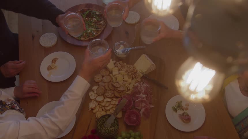 Friends clink glasses over a wooden table filled with crackers charcuterie and small plates under warm hanging lights