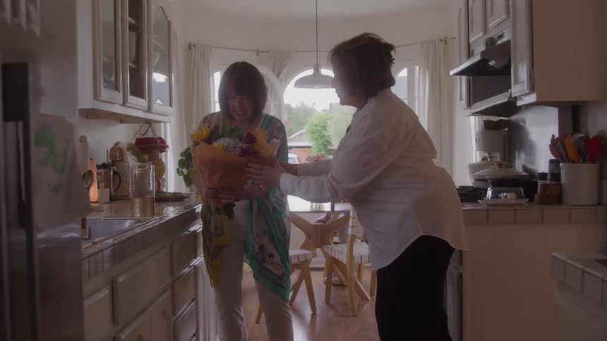 The video clip features two women talking in a bright kitchen with a third person seated at a dining table in the backgroundone woman holding a bouquet of flowers is smiling at the other suggesting a warm social visit or a gift being presented