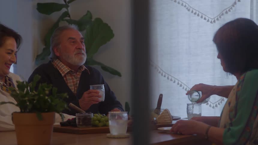 At a cozy table an older man smiles with a drink while another person pours water as they share snacks and conversation