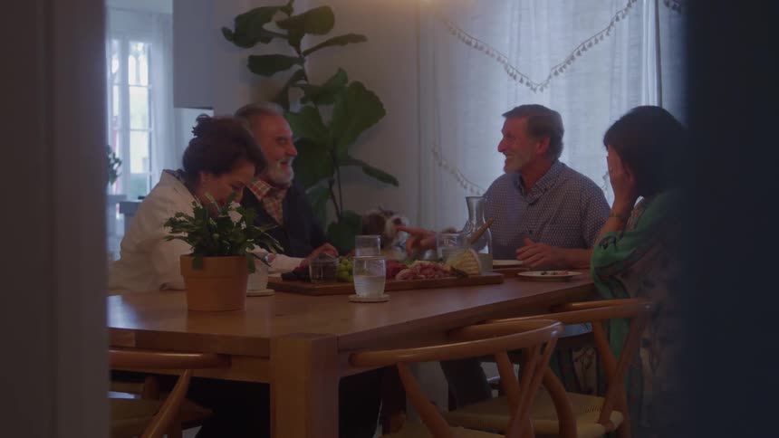 The video shows four adultstwo men and two womenseated around a large wooden dining table engaged in a conversation they are having a cheerful meal together with charcuterie and snacks visible on the table suggesting a social gathering