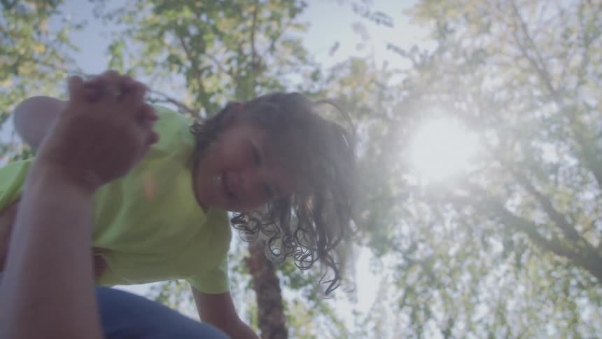 The video clip shows an adults arms holding a young child with curly hair up high against a bright sunlit backdrop of green trees and blue sky the child is wearing a bright lime green top and is being tossed playfully or held up in a celebr