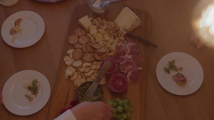 The video is a topdown slightly blurred closeup shot of a wooden serving board laden with various charcuterie items the board features a spread of cheeses crackers cured meats and a garnish like pesto indicating preparation for a shared meal or ap