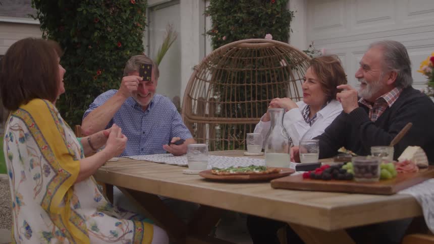 The video features four peopletwo men and two womenenjoying a sunny outdoor gathering around a wooden table they are having a cheerful conversation sharing snacks and one of the men is holding up a playing card in a seemingly playful moment