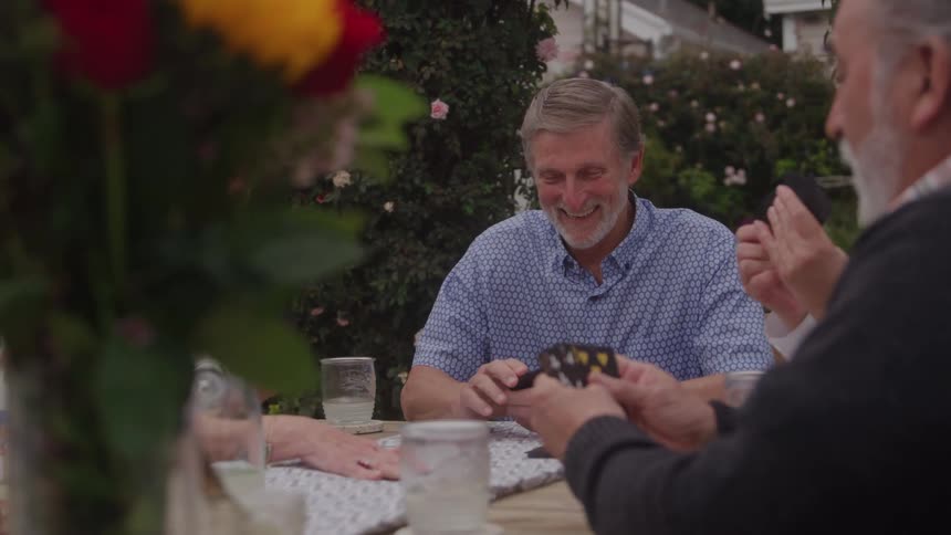 A group of friendstwo elderly men and two middleaged womensit together at an outdoor dining table under the midday sun laughing and enjoying a lively card game their faces light up with fun and excitement as they play