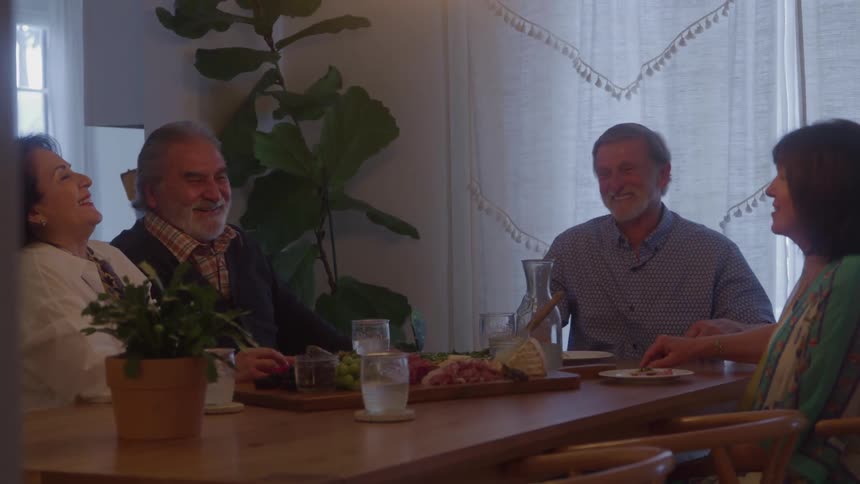 Four older people sit around a wooden dining table with a spread of food engaging in animated conversation and laughter with one man briefly raising his hand in the air during the discussion