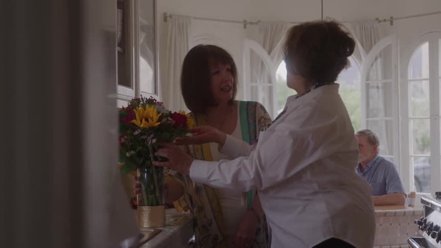 Two smiling middleaged women one in a white shirt and one in a printed shawl embrace next to a vase of sunflowers and red flowers in a bright kitchen with arched windows