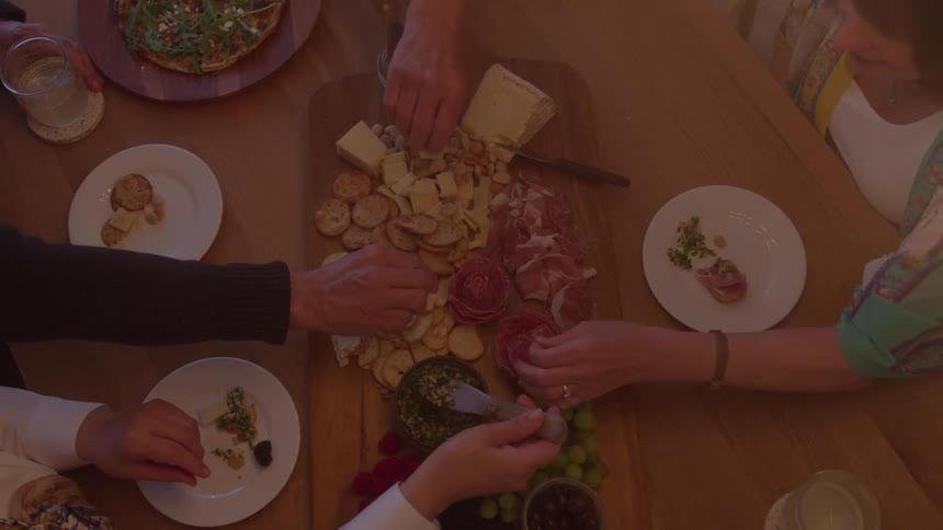 An overhead shot shows multiple hands reaching onto a wooden dining table to take items from a central wooden board covered with cheese crackers grapes and spreads with small white plates and drinking glasses around the board
