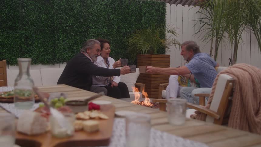 Two elderly couplesfeaturing two elderly men and their middleaged wivessit side by side in a cozy outdoor fireplace area with cheerful energy they toss their cups of coffee in a playful toast sharing laughter and warmth under the midday sun