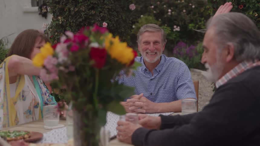 Four people are seated around a table outdoors surrounded by green foliage and a woman raises her hand to highfive another woman during a social gathering with a vase of flowers in the foreground